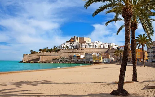 Küstenstadt mit Sandstrand, Palmen und historischem Schloss im Hintergrund.