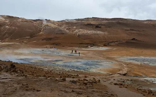 Menschen wandern durch eine karge, dampfende Landschaft mit braunen Hügeln im Hintergrund.