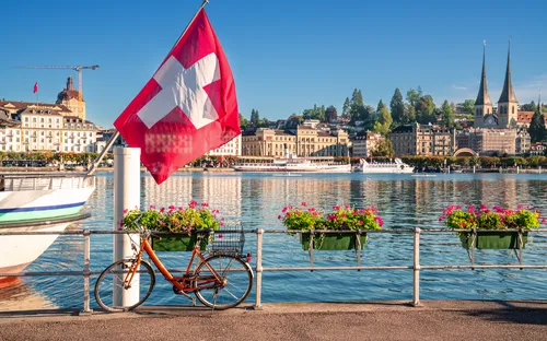 Schweizer Flagge, Fahrrad und blühende Blumen am Seeufer, Stadt im Hintergrund.