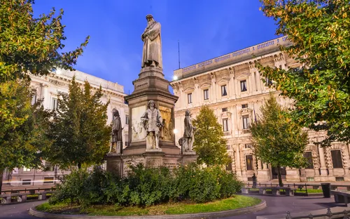 Denkmal auf einem Platz mit historischen Gebäuden im Hintergrund, umgeben von Bäumen.