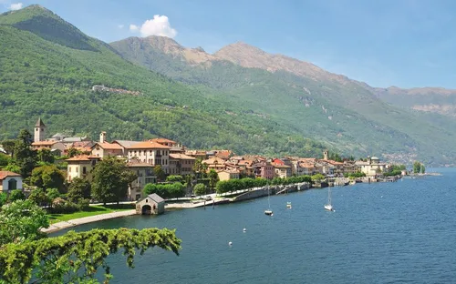 Stadt am See mit Alpen im Hintergrund, Bauten am Ufer, Boote im Wasser.