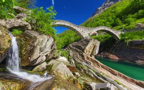 Steinbrücke über einen klaren Fluss mit Wasserfall in einer grünen Berglandschaft.