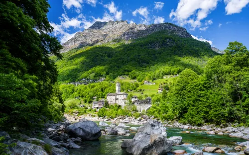 Bergdorf mit Kirche in grüner Landschaft und Fluss im Vordergrund unter blauem Himmel