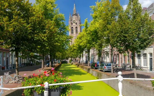Kanal in Delft mit Bäumen und Kirche im Hintergrund bei sonnigem Wetter.