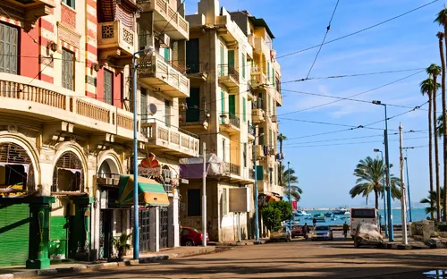 Straße mit alten Gebäuden und Palmen, im Hintergrund das Meer.