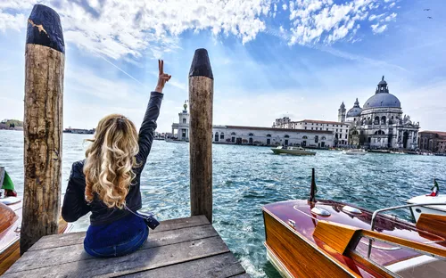 Frau auf Holzsteg macht ein Friedenszeichen in Venedig, italienische Architektur im Hintergrund.