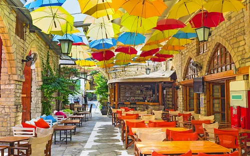 Bunte Regenschirme hängen über Terrasse eines Cafés mit Holztischen und Stühlen.