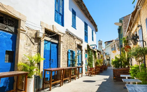 Gasse mit historischen Gebäuden, blauen Fensterläden und Außenterrasse bei Sonnenschein.