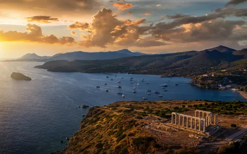 Küste bei Sonnenuntergang mit Tempel, Segelbooten und Bergen im Hintergrund.