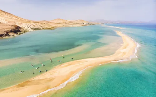 Luftaufnahme eines Strandes mit türkisblauem Wasser und Windsurfern.