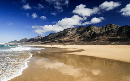 Sandstrand mit Bergen im Hintergrund unter blauem Himmel mit Wolken.