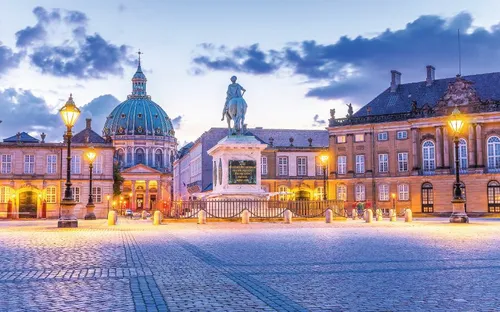 Historische Stadtansicht mit beleuchteter Kuppelkirche und Reiterstatue bei Dämmerung.
