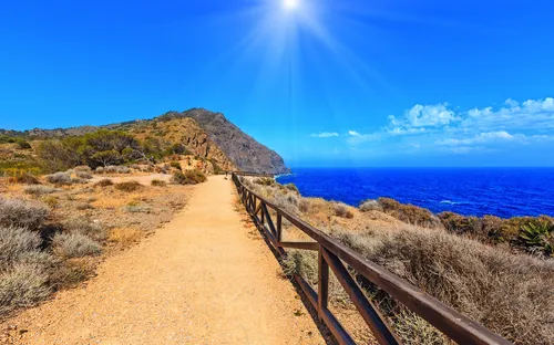 Wanderweg mit Geländer führt entlang der Küste bei strahlend blauem Himmel und Sonnenschein.