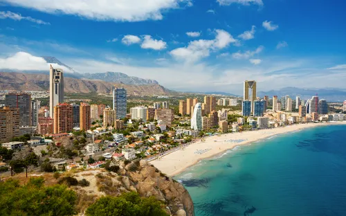 Küstenstadt mit Hochhäusern, Strand und Bergen im Hintergrund.