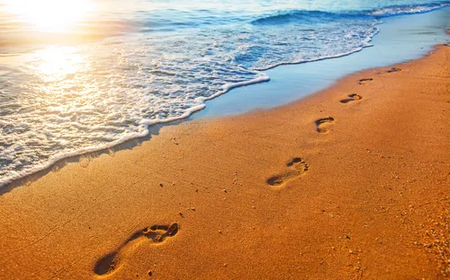 Fußspuren im Sand am Strand mit Meereswellen im Sonnenlicht.
