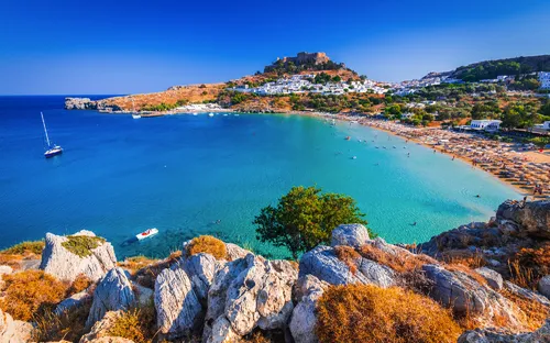 Küstenlandschaft mit blauem Meer, Segelboot und Sandstrand, im Hintergrund Hügel und Gebäude.
