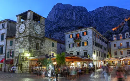 Abendlicher Platz mit Uhrturm und beleuchteten Gebäuden, Berge im Hintergrund.