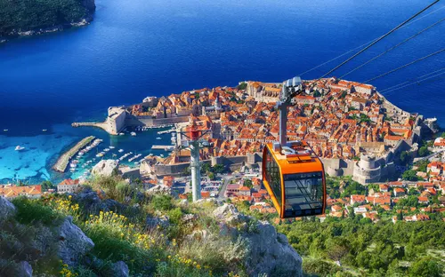Seilbahn überblickt historische Küstenstadt mit roter Ziegeldach-Skyline und tiefblauem Meer.