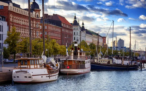 Boote im Hafen von Helsinki mit historischen Gebäuden und bewölktem Himmel im Hintergrund.