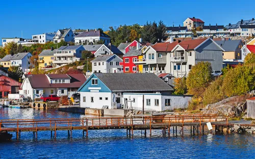 Bunte skandinavische Häuser am Wasser, umgeben von Bäumen und einer Pier im Vordergrund.