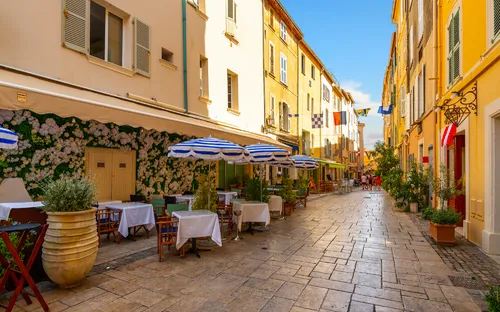 Eine sonnige Straße mit Cafétischen und bunten Gebäuden.