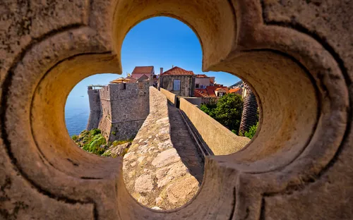 Blick durch steinernes Fenster auf historische Festungsmauer und Gebäude am Meer.