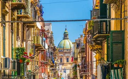 Schmale Straße in einer mediterranen Stadt mit Kirchturm im Hintergrund.