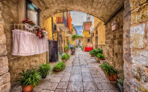 Steinbogen über Gasse mit Pflanzen und bunten Gebäuden in mediterranem Dorf.