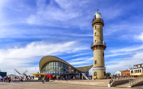 Leuchtturm mit modernem Gebäude und Menschen auf der Promenade bei sonnigem Wetter.