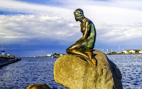 Skulptur einer Meerjungfrau auf einem Felsen am Wasser bei blauem Himmel.