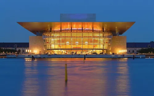 Ein beleuchtetes Gebäude mit modernem Design, das sich nachts am Wasser spiegelt.
