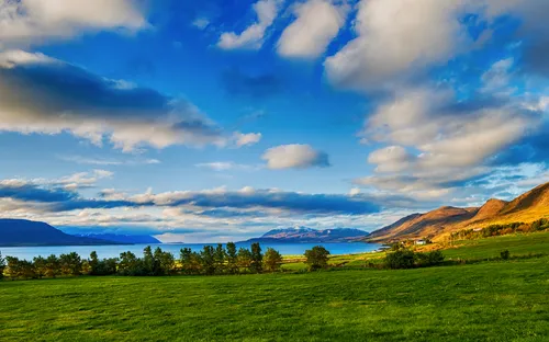 Grüne Wiese vor Bergen und See unter bewölktem Himmel