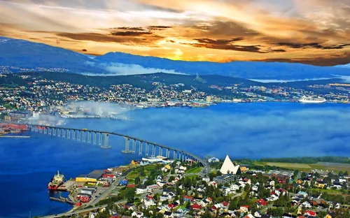 Stadtansicht mit Brücke, Häusern und Bergen bei Sonnenuntergang.