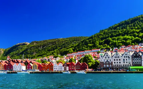 Häuserreihe am Wasser mit Bergen im Hintergrund bei klarem Himmel.
