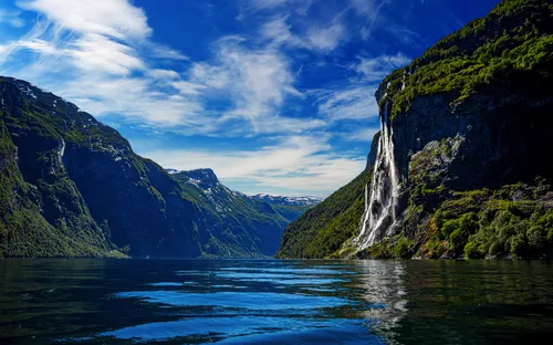 Fjord mit Wasserfall, umgeben von Bergen und blauem Himmel.
