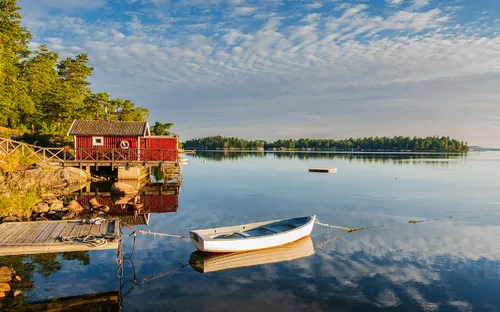 Boot am Steg vor rotem Haus an einem See bei klarem Himmel.