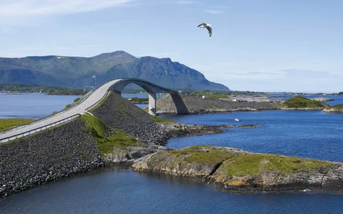 Brücke über blaues Wasser mit Bergen im Hintergrund und einem fliegenden Vogel.