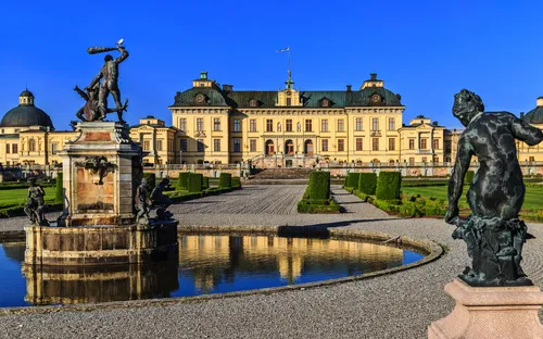 Schloss Drottningholm mit Gartenanlage und Statuen im Vordergrund an einem sonnigen Tag.