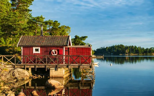 Rotes Holzhaus am Ufer eines Sees mit Bäumen im Hintergrund.