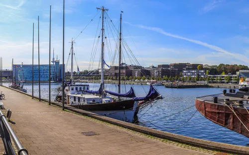 Hafenpromenade mit Segelbooten, blauem Himmel und Gebäuden im Hintergrund.