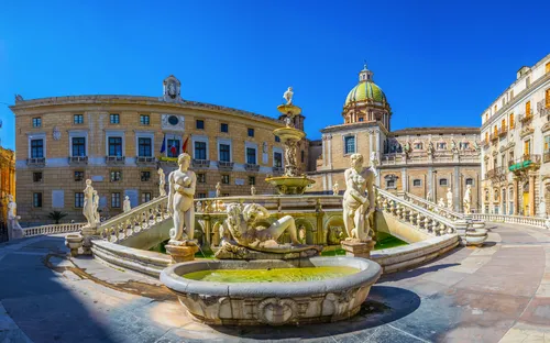 Historischer Brunnen mit Statuen vor alten Gebäuden und blauen Himmel.