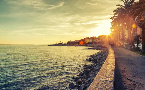 Sonnenuntergang an einer Küstenpromenade mit Palmen, Meer und einer felsigen Küste.