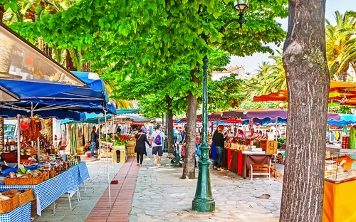 Marktstand unter Bäumen mit Besuchern und bunten Schirmen.