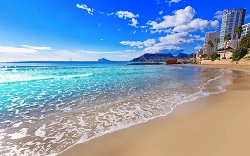 Strand mit klarem Wasser, blauem Himmel und einigen Wolken, im Hintergrund Gebäude und Berge.