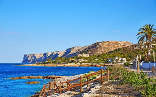 Küste mit blauem Meer, Felsen und Palmen im Sonnenschein.