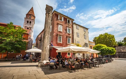 Menschen in einem Café im Freien auf einem sonnigen Platz mit historischen Gebäuden.