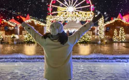 Person mit Winterkleidung schaut auf Weihnachtsmarkt im Schnee.