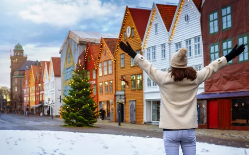Person mit Winterbekleidung vor bunten Holzhäusern und Tannenbaum im Schnee.
