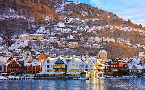 Winterlandschaft mit bunten Häusern am Wasser und schneebedeckten Hügeln im Hintergrund.